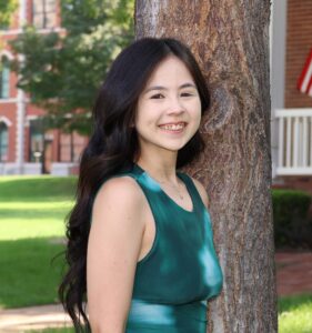 Faith Oshiro, Miss Northwestern contestant, in a green sleeveless top, standing beside a tree with a brick building behind her.