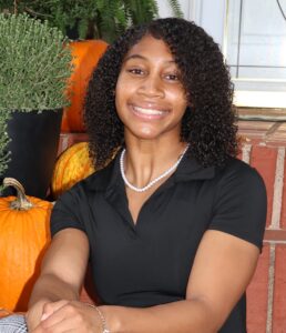 Sierra Brooks, Miss Northwestern contestant, in a black polo shirt and pearl necklace, seated in front of pumpkins and a brick building.