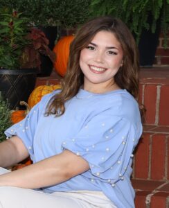 Abigail Miller, Miss Northwestern contestant, in a light blue pearl-trimmed top, seated on brick steps with pumpkins and plants.