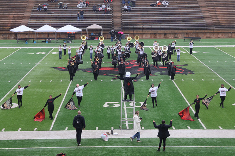 The Northwestern Oklahoma State University Ranger marching band performs on Ranger Field around the midfield Ranger logo, with the flag line spread across the 50-yard line and a drum major conducting from a ladder, as fans watch from the stands on an overcast day.