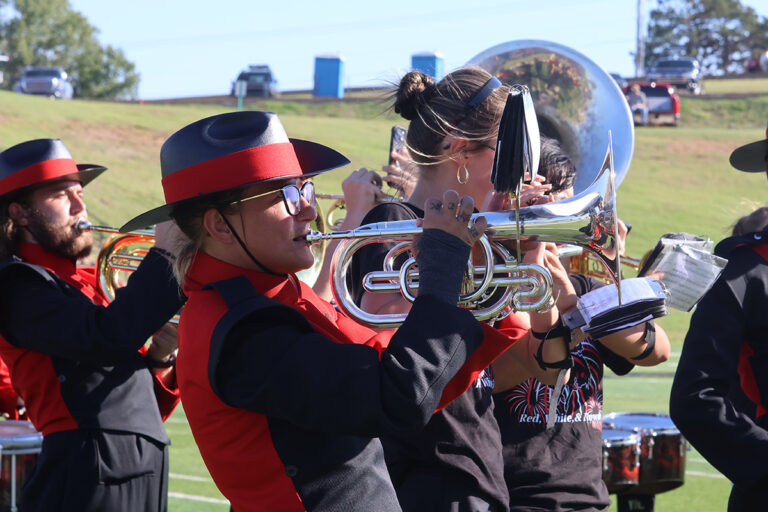 Ranger Marching Band members in black and red uniforms and brimmed hats play brass instruments and saxophone during halftime of the Northwestern Oklahoma State University homecoming football game.