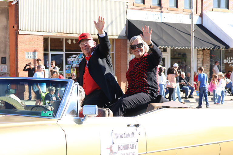 Parade marshal Verne Metcalf and his wife, Gloria, smile and wave from the back of a gold convertible during the Northwestern Oklahoma State University homecoming parade in downtown Alva, Oklahoma. A sign on the car door names Verne Metcalf as honorary parade marshal.