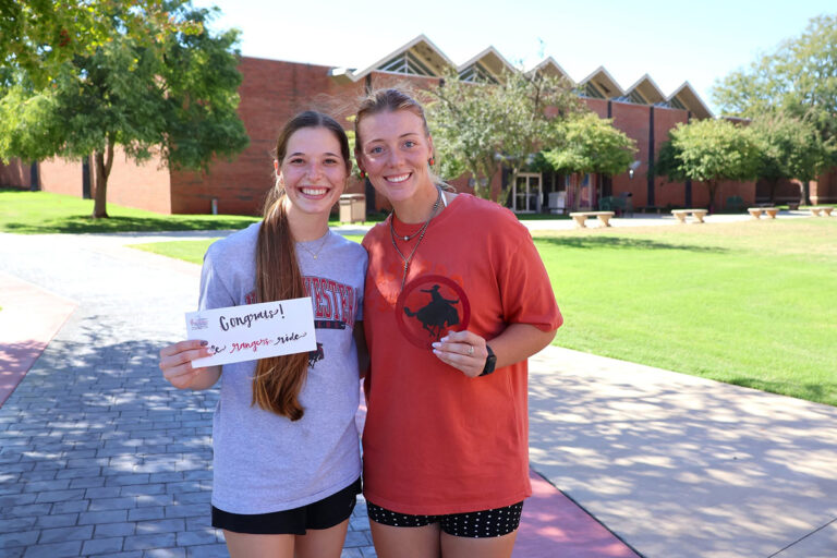 Medallion hunt winners Haylee Durrence and Aspen Loafman smile on a Northwestern Oklahoma State University campus sidewalk; Durrence holds a handwritten note reading “Congrats! Ride Rangers Ride” and Loafman holds a medallion with a bucking horse and rider silhouette.