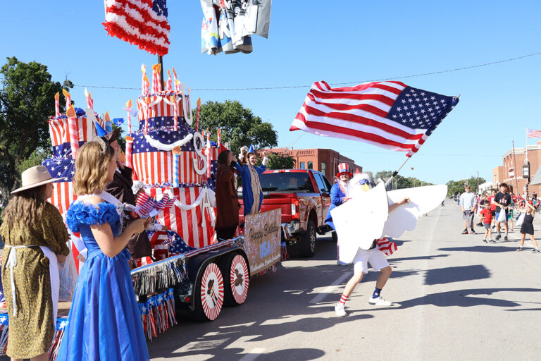 A person in an eagle costume waves a large American flag beside the Northwestern Scholar Ambassadors float, a red, white and blue birthday cake spelling “USA” with a “Happy Birthday USA” sign, as riders in colonial and patriotic costumes ride along during the Northwestern Oklahoma State University homecoming parade in Alva, Oklahoma.