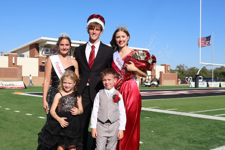 Ranger King Coy Bullard, wearing a crown, stands on Ranger Field with Ranger Queen Alaina Pierce and Freshman Queen Chandler Ogden, with flower girl Ashton Joy Mosburg and crown bearer Branch Allen Anderson in front, at Northwestern Oklahoma State University.