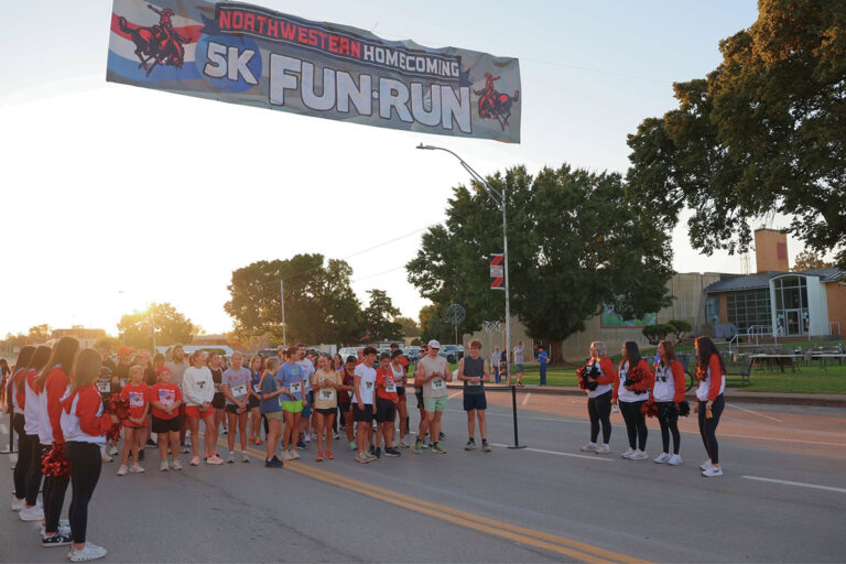 Runners wearing race bibs gather at the start line under a banner reading “Northwestern Homecoming 5K Fun-Run” as Northwestern cheerleaders with red pompoms line both sides of the street at sunrise in Alva, Oklahoma.