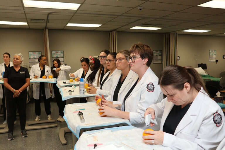 Doctoral nursing students in white lab coats work at draped instrument tables in the Northwestern Oklahoma State University simulation lab, practicing procedure skills on oranges with surgical instruments while two faculty members in black scrubs observe.