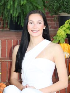 Raley Hooper, Miss Northwestern contestant, in a white one-shoulder outfit, seated on brick steps with pumpkins and ferns.