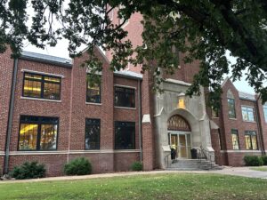 Front entrance of Herod Hall at Northwestern Oklahoma State University in Alva, Oklahoma. The two-story red brick building has a carved limestone archway with the word “Administration” above glass doors, framed by tree branches.