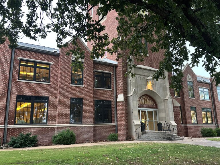 Front entrance of Herod Hall at Northwestern Oklahoma State University in Alva, Oklahoma. The two-story red brick building has a carved limestone archway with the word “Administration” above glass doors, framed by tree branches.