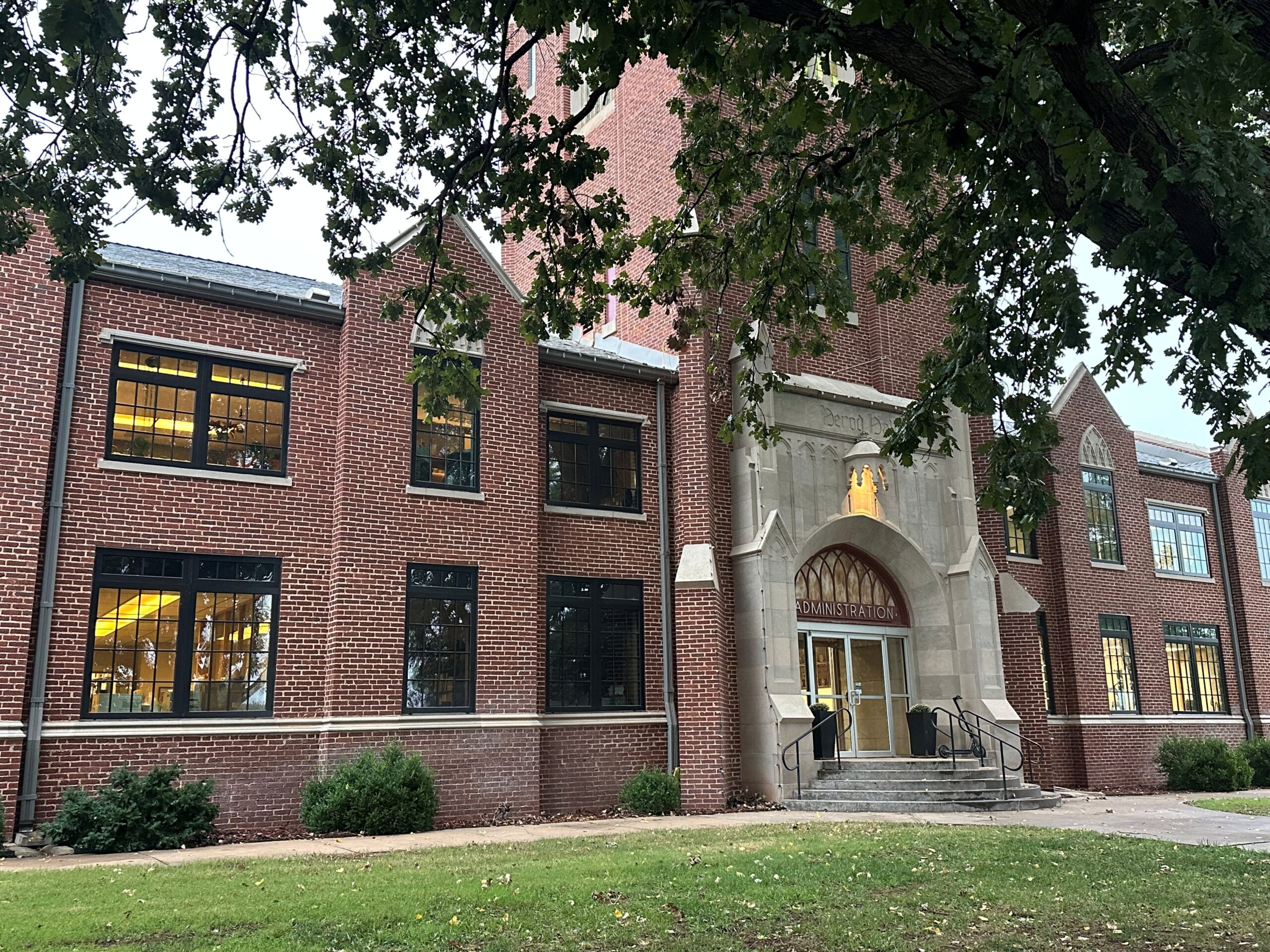 Front entrance of Herod Hall at Northwestern Oklahoma State University in Alva, Oklahoma. The two-story red brick building has a carved limestone archway with the word “Administration” above glass doors, framed by tree branches.