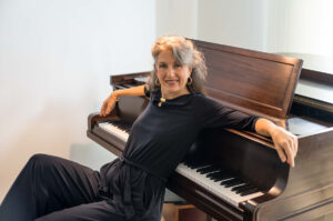 Jazz vocalist Alexis Cole sits on a bench beside a dark wood grand piano, turned toward the camera and smiling. She wears a black long-sleeved top and trousers with gold hoop earrings and a gold bracelet. One arm rests along the top of the piano and her other hand rests near the keys. Sheet music sits on the music desk, and the background is a plain white wall in soft daylight.