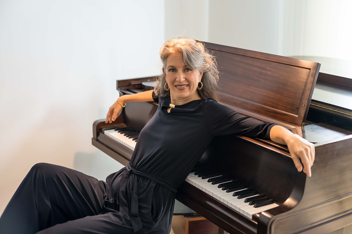 Jazz vocalist Alexis Cole sits on a bench beside a dark wood grand piano, turned toward the camera and smiling. She wears a black long-sleeved top and trousers with gold hoop earrings and a gold bracelet. One arm rests along the top of the piano and her other hand rests near the keys. Sheet music sits on the music desk, and the background is a plain white wall in soft daylight.