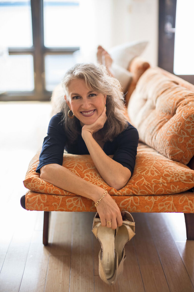 Jazz vocalist Alexis Cole lies on her front across an orange floral upholstered bench, propped on her forearms and resting her chin on one hand as she smiles at the camera. She wears a navy three-quarter-sleeve top, gold hoop earrings and a gold chain bracelet, and holds a pair of tan strappy heels in one hand. Her bare feet are raised behind her. Behind the bench are a cream cushion and a bright window; the floor is dark hardwood.