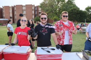 Hadley Bohlen, Brayden Stoner and Austin Rankin stand behind red insulated food carriers, wearing serving gloves and flower leis, as they hand out free food to students on the lawn during the 2025 Aloha! Northwestern event at Northwestern Oklahoma State University. A brick residence hall and students at picnic tables are in the background.