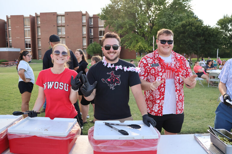 Hadley Bohlen, Brayden Stoner and Austin Rankin stand behind red insulated food carriers, wearing serving gloves and flower leis, as they hand out free food to students on the lawn during the 2025 Aloha! Northwestern event at Northwestern Oklahoma State University. A brick residence hall and students at picnic tables are in the background.