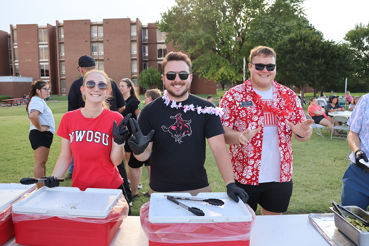 Hadley Bohlen, Brayden Stoner and Austin Rankin stand behind red insulated food carriers, wearing serving gloves and flower leis, as they hand out free food to students on the lawn during the 2025 Aloha! Northwestern event at Northwestern Oklahoma State University. A brick residence hall and students at picnic tables are in the background.