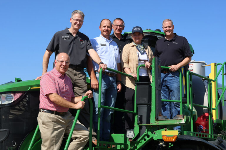 Six men and women stand on the platform and steps of a green John Deere sprayer under a clear blue sky. From left: Jeff Hickman of the Oklahoma Grain and Feed Association, Dr. Steve Maier of Northwestern Oklahoma State University, Dr. Kent Martin of Northwestern, Bob Baker of 4 Generations Organic, Dr. Karen St. Germain of NASA and Jim Bridenstine of Quantum Space.