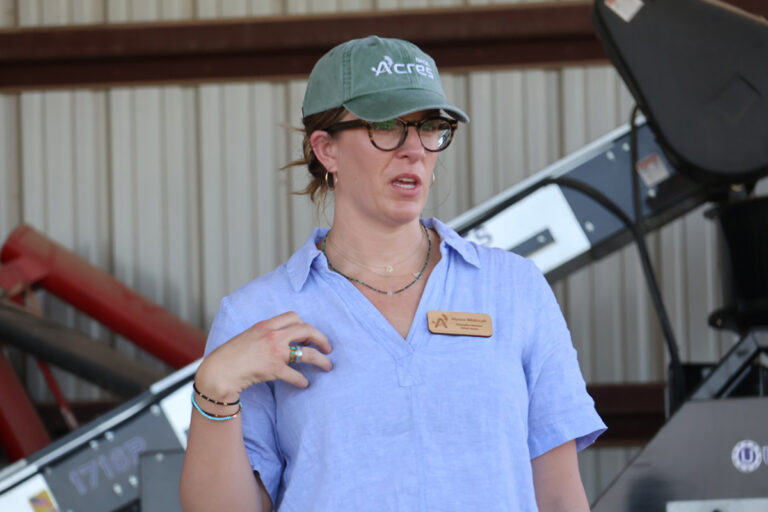 Dr. Alyssa Whitcraft, executive director of NASA Acres, speaks beside seed-cleaning equipment inside the shop at Bush Seed Company, wearing a NASA Acres cap and a name badge reading Executive Director, NASA Acres.