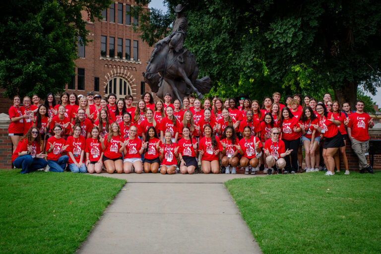 The 2025 Camp Ranger group of incoming Northwestern Oklahoma State University freshmen, all wearing red Camp Ranger T-shirts, poses in rows on the lawn in front of a bronze statue of a cowboy riding a bucking horse on the Alva campus.
