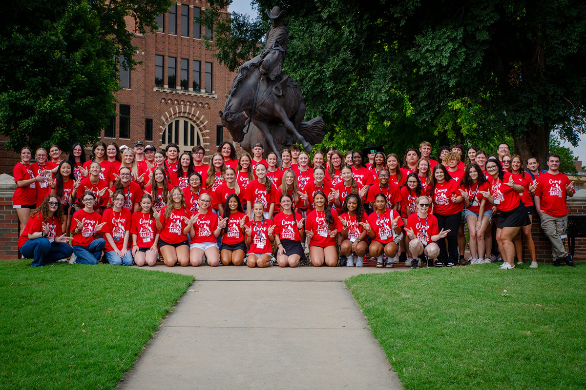 The 2025 Camp Ranger group of incoming Northwestern Oklahoma State University freshmen, all wearing red Camp Ranger T-shirts, poses in rows on the lawn in front of a bronze statue of a cowboy riding a bucking horse on the Alva campus.