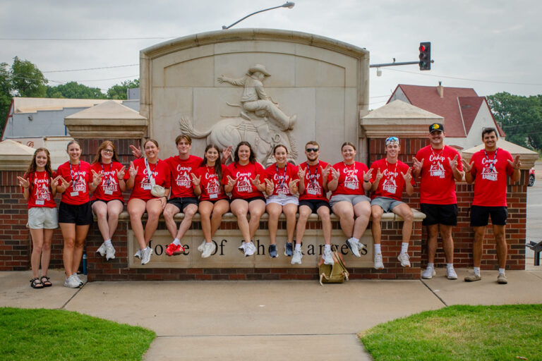 Thirteen members of the 2025 Northwestern Scholar Ambassadors in red Camp Ranger T-shirts make the Ride, Rangers, Ride hand sign while seated and standing on the Home of the Rangers monument in Alva.