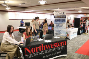 Wide view of last year’s Northwestern Oklahoma State University Career and Internship Fair. In the foreground, three women work on laptops at a black table with a banner reading Northwestern Oklahoma State University Graduate Studies. Behind them, attendees visit employer tables, including a banner reading Kiowa District Healthcare, Excellence Is Our Standard, and tables for Oklahoma Human Services and Blue Sky Crop Consulting. A uniformed officer stands at a table in the back.