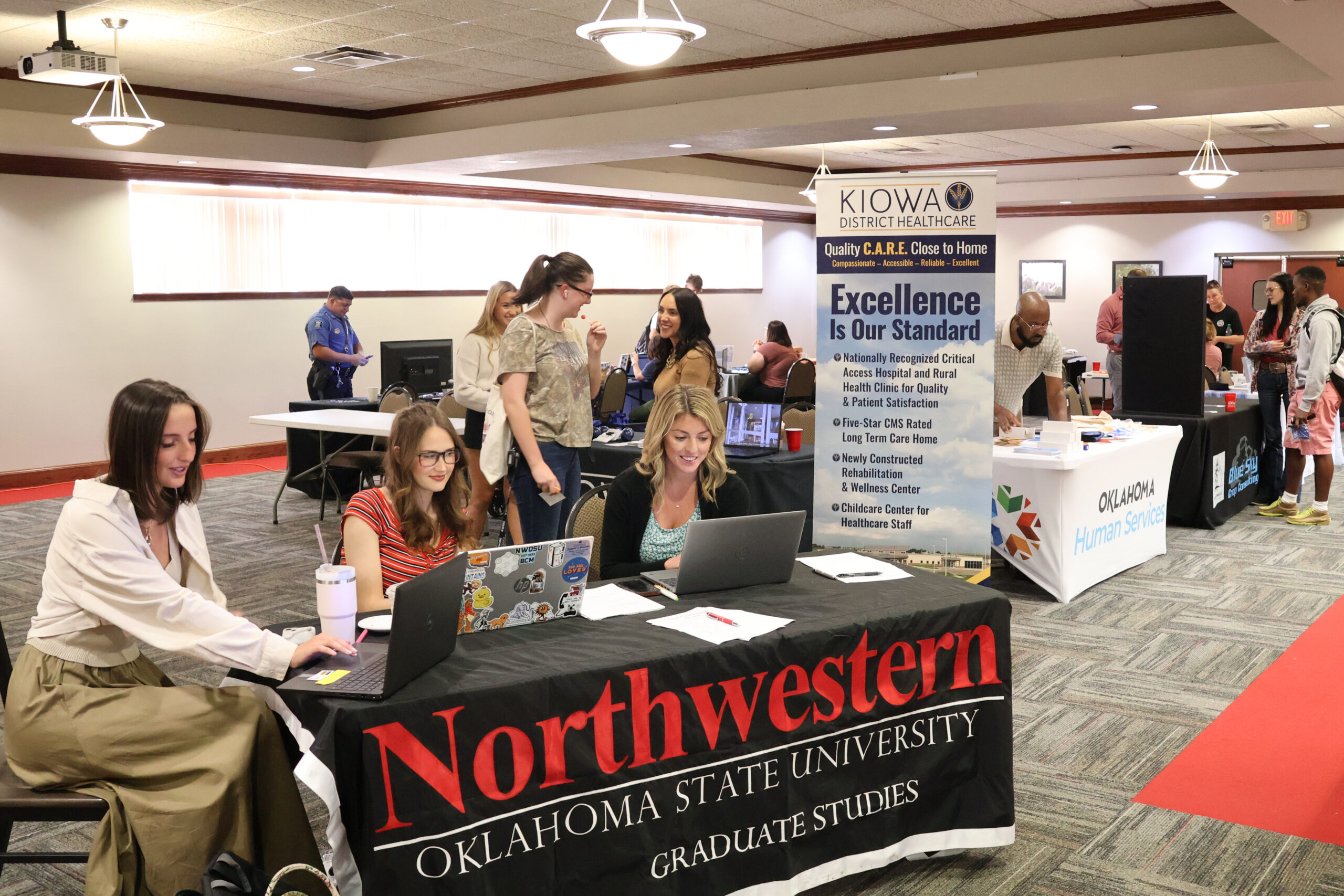 Wide view of last year’s Northwestern Oklahoma State University Career and Internship Fair. In the foreground, three women work on laptops at a black table with a banner reading Northwestern Oklahoma State University Graduate Studies. Behind them, attendees visit employer tables, including a banner reading Kiowa District Healthcare, Excellence Is Our Standard, and tables for Oklahoma Human Services and Blue Sky Crop Consulting. A uniformed officer stands at a table in the back.