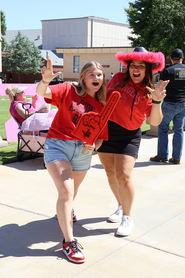 Two Northwestern Oklahoma State University students in red Rangers shirts pose on a campus sidewalk during the 2025 Howdy Week clubs and organizations fair. One holds a red foam finger reading Go Rangers; the other wears a bright pink cowboy hat. Organization booths and tables are set up behind them.