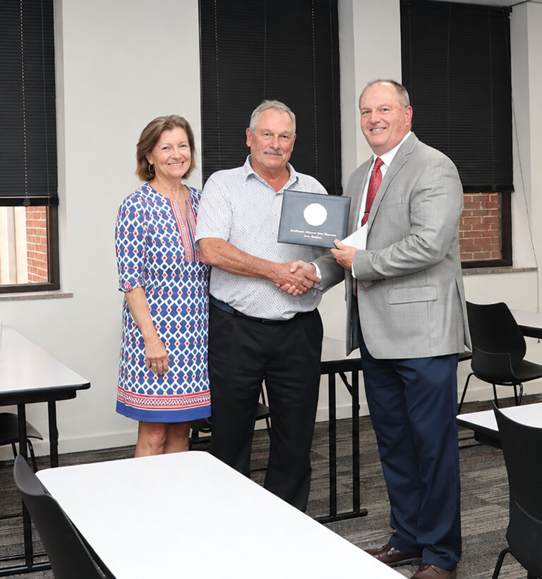 Dr. Bo Hannaford, right, presents Dave Johnson, center, with a diploma cover for his honorary Doctorate of Science and Business Leadership as the two shake hands. Johnson's wife, Louise, stands at left in a blue, red and white patterned dress. All three smile in a classroom with white walls, black window blinds, white tables and black chairs.