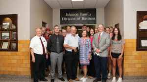 Business faculty, members of the Johnson family and students stand together in a hallway beneath a black sign with white letters reading "Johnson Family Division of Business." A man near the center of the front row holds a black diploma cover with a gold seal. Yellow tile lines the lower walls, with a wooden display case of framed black-and-white photographs at left and framed certificates at right.