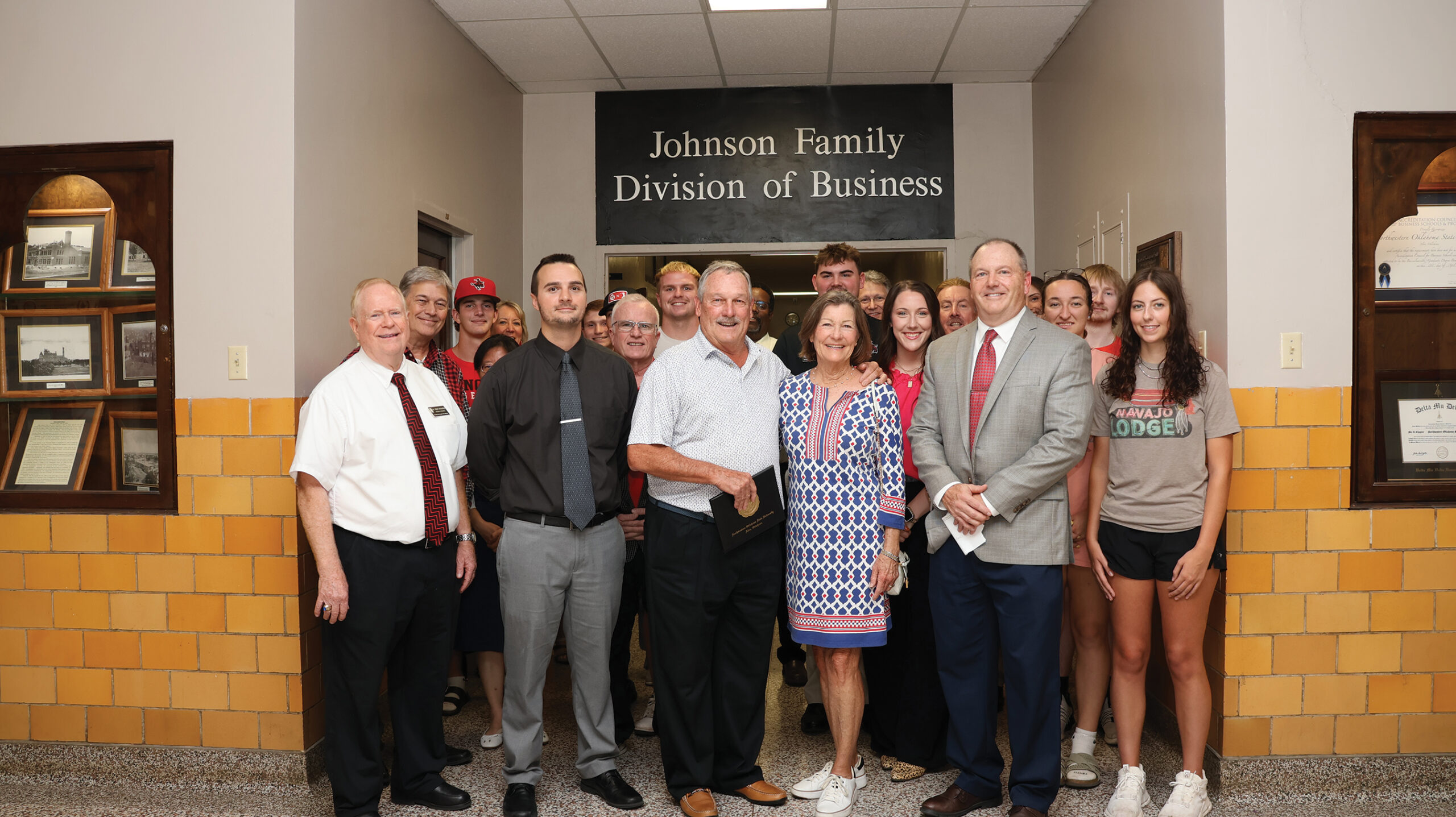 Business faculty, members of the Johnson family and students stand together in a hallway beneath a black sign with white letters reading "Johnson Family Division of Business." A man near the center of the front row holds a black diploma cover with a gold seal. Yellow tile lines the lower walls, with a wooden display case of framed black-and-white photographs at left and framed certificates at right.
