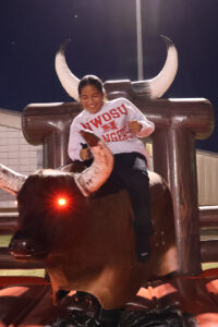 A smiling Northwestern Oklahoma State University student in a white NWOSU Rangers sweatshirt grips the handle and rides an inflatable mechanical bull with a glowing red eye at night, lit by a stadium light pole, during 2025 Student Government Association homecoming activities.