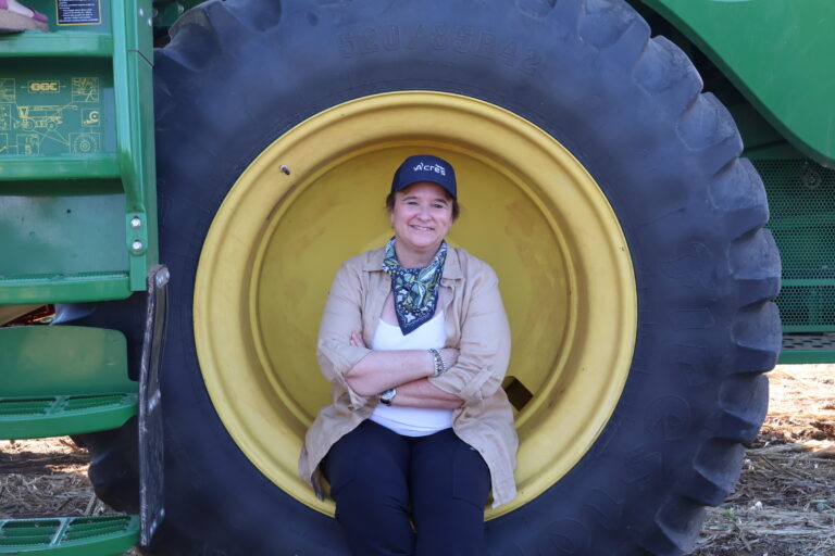 Dr. Karen St. Germain, director of NASA's Earth Science Division, sits smiling with her arms crossed inside the yellow wheel hub of a green John Deere combine, wearing a NASA Acres cap and a patterned bandana.