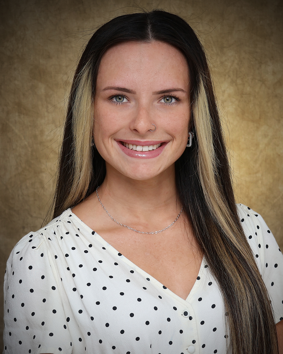 Cadence Long-Torres, a Northwestern Oklahoma State University senior psychology major, smiles in a portrait wearing a white blouse with black polka dots against a mottled gold backdrop.