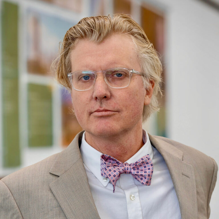 Dr. Mark J. Sundahl, professor of law and director of the Global Space Law Center at Cleveland State University College of Law, wearing clear-framed glasses, a tan suit jacket, a white button-down shirt and a pink patterned bow tie, in front of a softly blurred gallery wall hung with framed artwork.