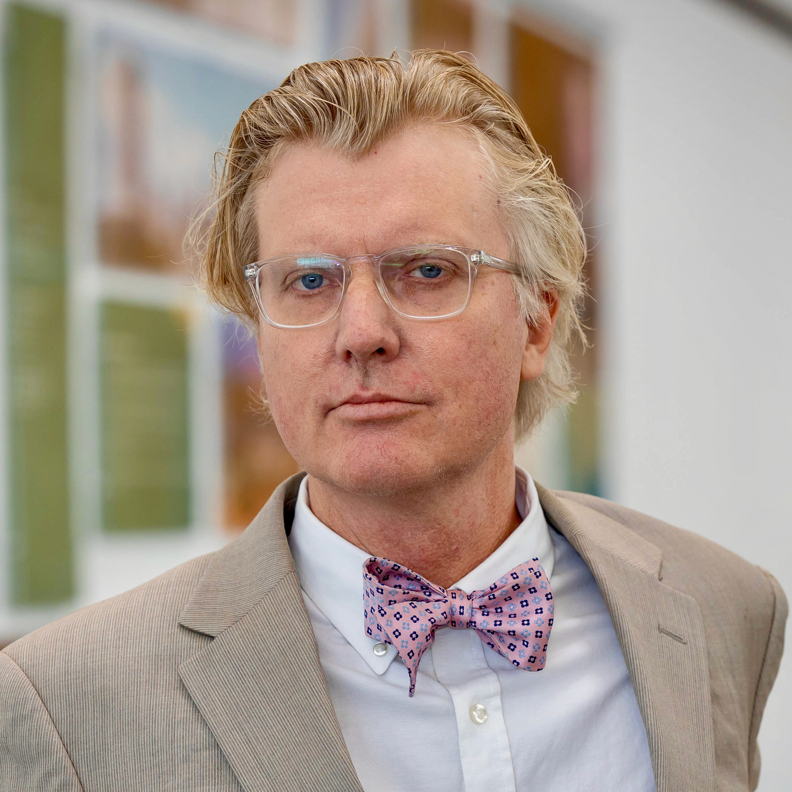 Dr. Mark J. Sundahl, professor of law and director of the Global Space Law Center at Cleveland State University College of Law, wearing clear-framed glasses, a tan suit jacket, a white button-down shirt and a pink patterned bow tie, in front of a softly blurred gallery wall hung with framed artwork.