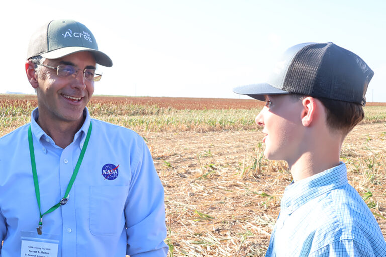 Forrest Melton, senior research scientist at NASA Ames Research Center, wearing a NASA-branded shirt and a lanyard badge reading NASA Listening Tour 2026, smiles while talking with a young man in a plaid shirt and cap at the edge of a harvested milo field.