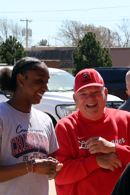 Verne Metcalf (right), in a red cap and red sweatshirt, and Kira Bass (left), in a gray Great American Conference champions T-shirt, laugh together outdoors at Lady Ranger Day