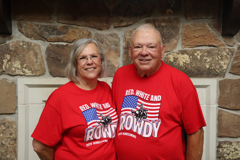 Verne Metcalf (right), 2026 Northwestern homecoming parade marshal, and his wife, Gloria (left), smile in matching red “Red, White and Rowdy” 2026 Homecoming T-shirts in front of a stone fireplace at the Homestead in Alva, Oklahoma
