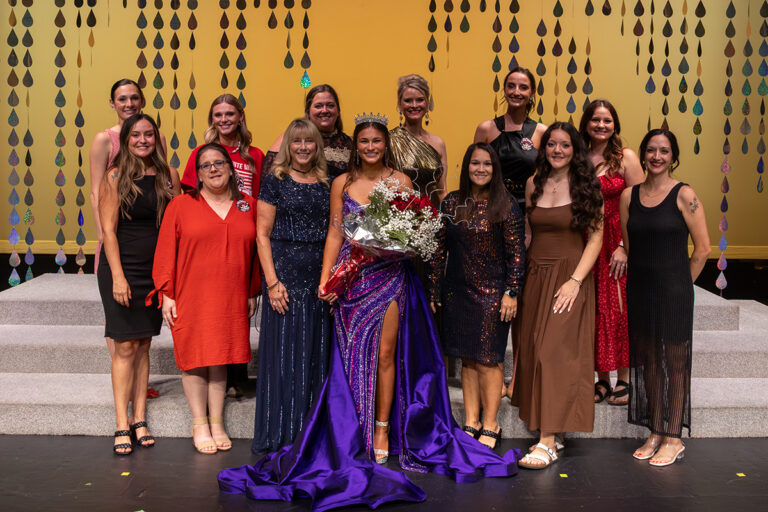 Miss Cinderella 2026 Kaycie Shelton, wearing a crown and a purple sequined gown and holding red roses, stands at the center of a group of women from the Miss Cinderella Pageant committee on stage at Northwestern Oklahoma State University's 74th Miss Cinderella Pageant in Alva, Oklahoma. All smile at the camera in front of a gold backdrop hung with iridescent teardrop strands.