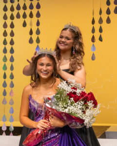 Miss Cinderella 2025 Madeline Ryerson, wearing a tiara and a black gown, stands behind Kaycie Shelton and places a crown on her head on stage at Northwestern Oklahoma State University's 74th Miss Cinderella Pageant in Alva, Oklahoma. Shelton, Miss Cinderella 2026, smiles in a purple sequined gown and holds a bouquet of red roses and baby's breath. Strands of iridescent teardrop shapes hang against a gold backdrop behind them.