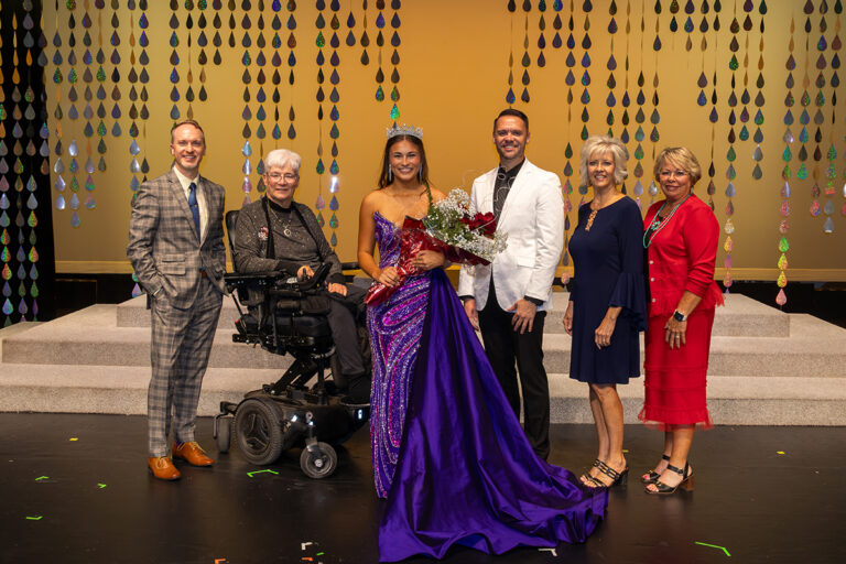 Miss Cinderella 2026 Kaycie Shelton, wearing a crown and a purple sequined gown and holding red roses, stands on stage with the judges of Northwestern Oklahoma State University's 74th Miss Cinderella Pageant in Alva, Oklahoma. Left to right: Brandt Sterling, in a gray plaid suit; Elda Jones, seated; Shelton; Kody Humphrey, in a white jacket; Myra Davison, in navy; and Paula McMurphy, in red. All smile at the camera in front of a gold backdrop.