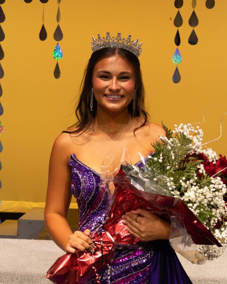 Kaycie Shelton, Miss Cinderella 2026, smiles at the camera on stage at Northwestern Oklahoma State University's 74th Miss Cinderella Pageant in Alva, Oklahoma, wearing a crown and a purple sequined gown and holding a bouquet of red roses and baby's breath wrapped in red. A gold backdrop hung with iridescent teardrop strands is behind her.