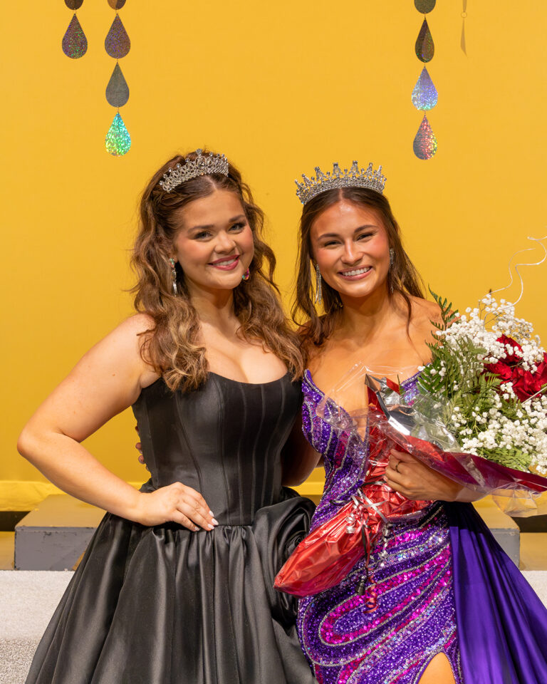 Miss Cinderella 2025 Madeline Ryerson, in a tiara and black gown, stands beside Miss Cinderella 2026 Kaycie Shelton on stage at Northwestern Oklahoma State University's 74th Miss Cinderella Pageant in Alva, Oklahoma. Shelton wears a crown and a purple sequined gown and holds a bouquet of red roses and baby's breath. Both smile at the camera in front of a gold backdrop hung with iridescent teardrops.
