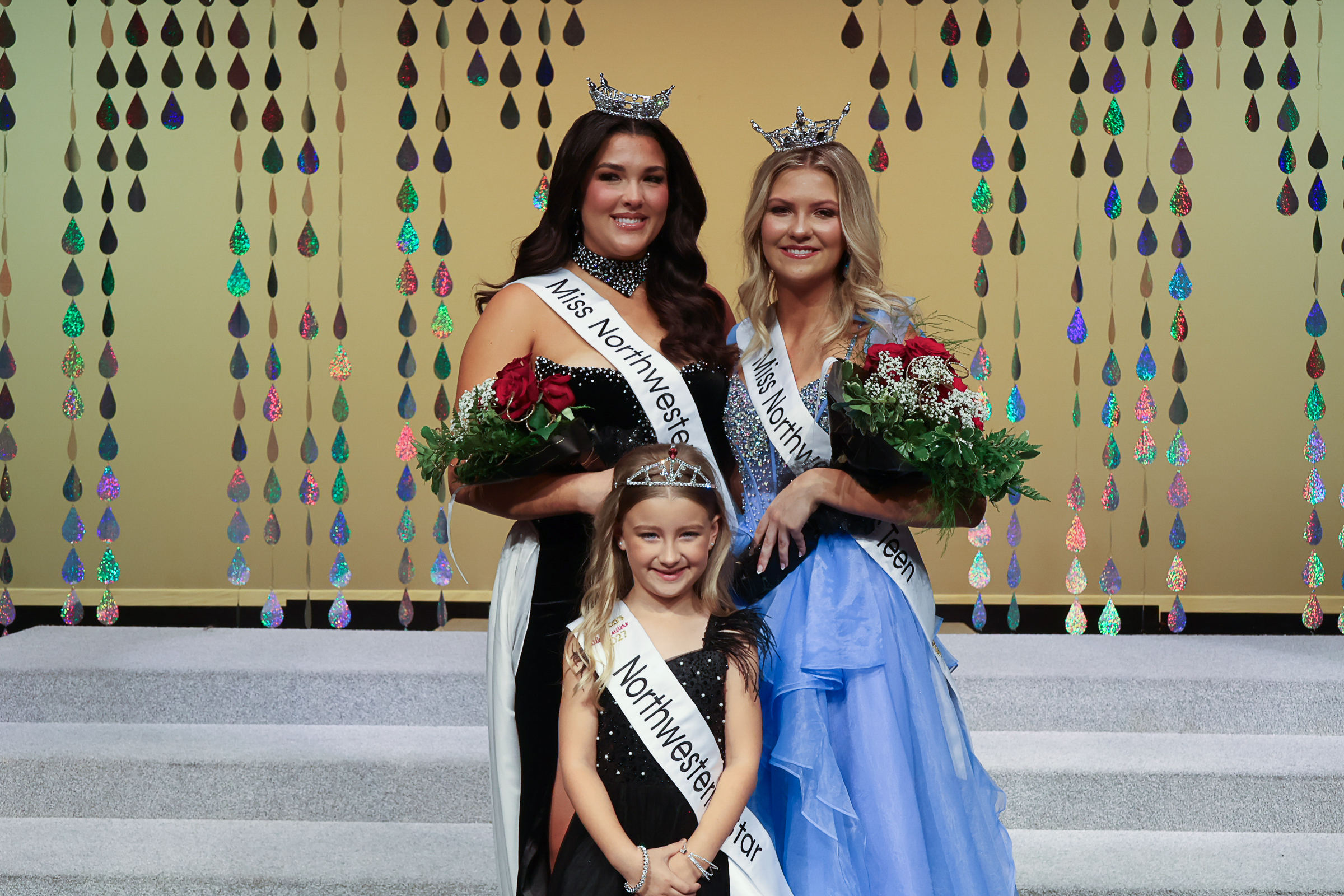 Raegan Bleything, Miss Northwestern 2027, and Nelley Burnett, Miss Northwestern’s Teen 2027, both wearing crowns and sashes and holding red roses, stand on stage in Fellers Family Auditorium in front of a gold backdrop marked 60. Northwestern Star 2027 Harper Higgins, in a black dress, tiara and Northwestern Star sash, stands in front of them.