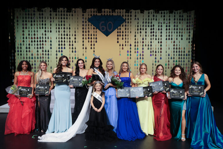 Eleven 2027 Miss Northwestern and Miss Northwestern’s Teen winners in evening gowns stand in a row on stage in Fellers Family Auditorium, most holding award certificates, in front of a gold backdrop marked 60. Miss Northwestern 2027 Raegan Bleything and Miss Northwestern’s Teen 2027 Nelley Burnett stand in the center wearing crowns and sashes and holding red roses, with Northwestern Star 2027 Harper Higgins in front of them. Left to right: Sierra Brooks, Trinity Tuell, Jayla Adams, Raley Hooper, Bleything, Burnett, Kirstin Fennema, Olivia Church, Gianna Latta, Rebecka “Becky” Carlson and Loralei Welch.