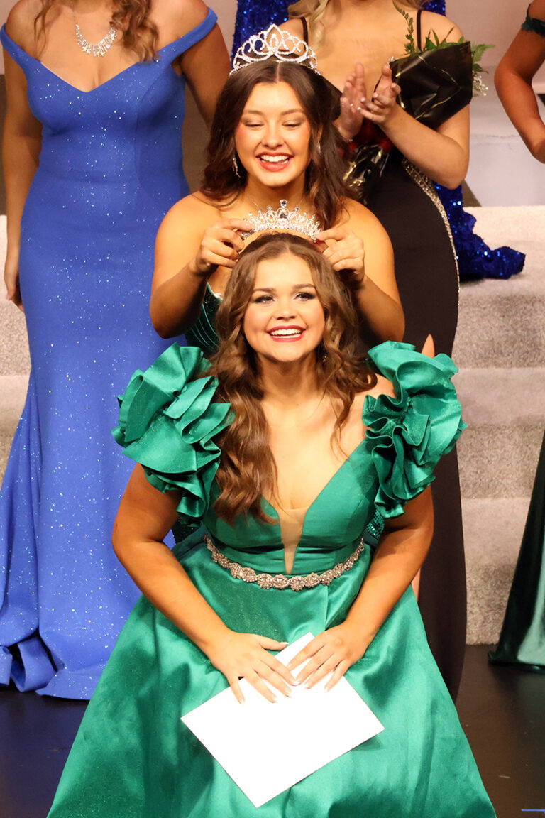 Madeline Ryerson, 2025 Miss Cinderella, seated in an emerald satin gown with ruffled sleeves and holding a white envelope, smiles as Zoey Roberts, 2024 Miss Cinderella, standing behind her in a crown, places a tiara on her head. Other contestants stand on the steps behind them.