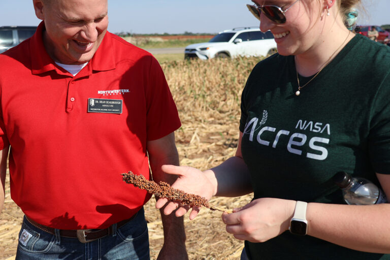 Dr. Dean Scarbrough, chair of the agriculture department at Northwestern Oklahoma State University, in a red Northwestern Rangers polo with a name badge, laughs while holding a milo head in his open palm alongside Faith Riensche in a dark green NASA Acres T-shirt, standing in a harvested milo field.
