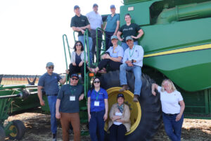 Twelve NASA and NASA Acres staff members pose on and around a green John Deere combine in a harvested field. Several wear NASA Acres caps and shirts.